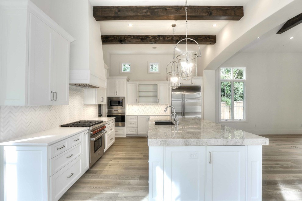 Modern inredning av ett kök och matrum, med en undermonterad diskho, vita skåp, rostfria vitvaror, en köksö, marmorbänkskiva, vitt stänkskydd, stänkskydd i glaskakel och ljust trägolv