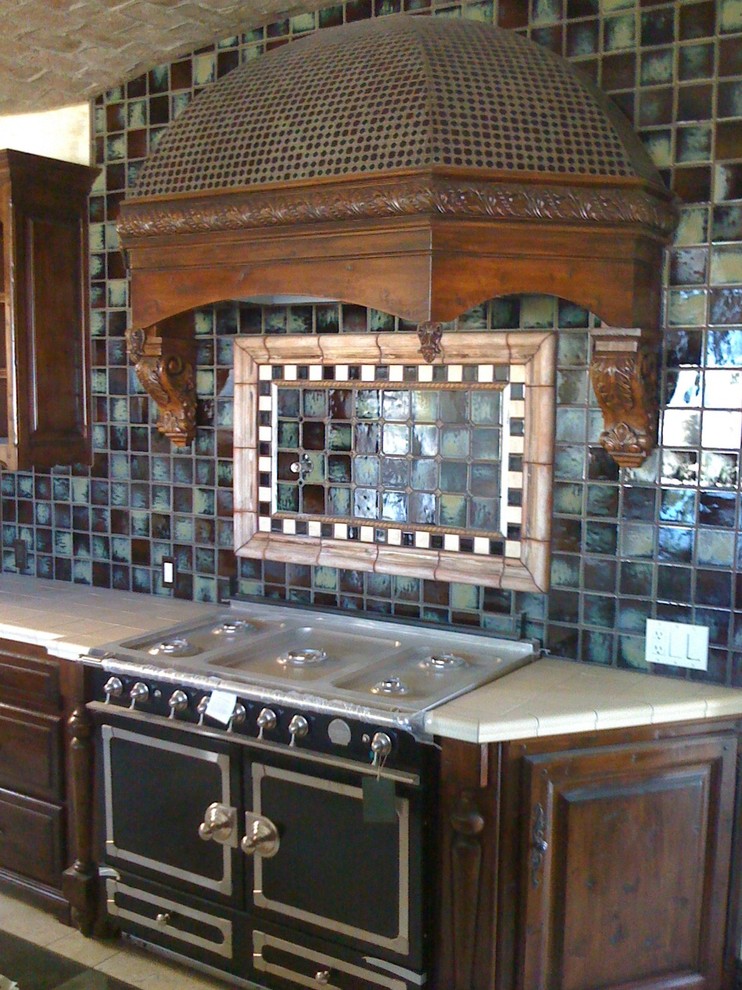 Immagine di una cucina lineare mediterranea di medie dimensioni con ante con bugna sagomata, ante in legno bruno, top piastrellato, paraspruzzi multicolore, paraspruzzi con piastrelle a mosaico, elettrodomestici neri e pavimento in travertino