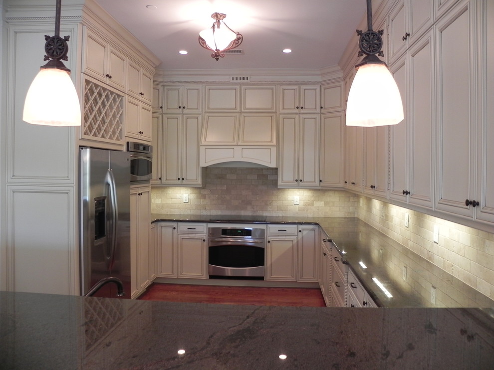 Example of a mid-sized classic u-shaped medium tone wood floor and brown floor enclosed kitchen design in Charlotte with recessed-panel cabinets, white cabinets, stainless steel appliances, a peninsula, granite countertops, beige backsplash and stone tile backsplash