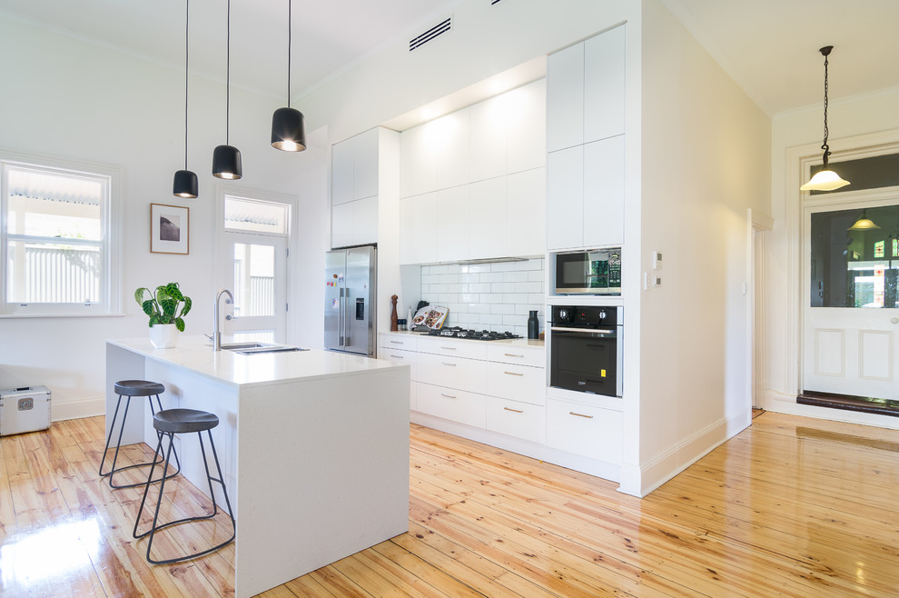 Photo of a large modern galley kitchen/diner in Auckland with a submerged sink, white cabinets, composite countertops, white splashback, ceramic splashback, white appliances, medium hardwood flooring, an island, beige floors and white worktops.