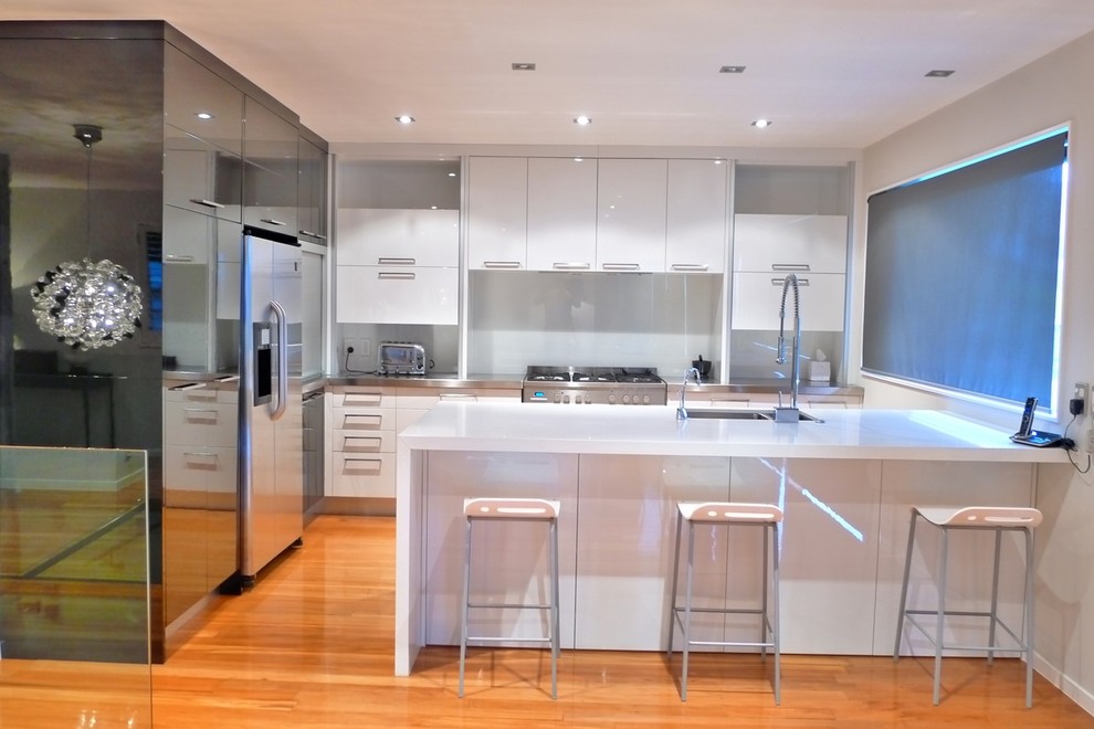 Inspiration for a mid-sized modern l-shaped medium tone wood floor kitchen remodel in Auckland with a double-bowl sink, flat-panel cabinets, white backsplash, glass sheet backsplash, stainless steel appliances and a peninsula
