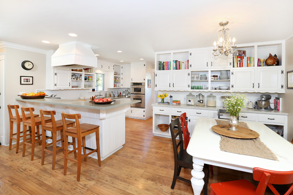 This is an example of a farmhouse l-shaped kitchen/diner in Orange County with raised-panel cabinets, white cabinets, wood worktops, white splashback, white appliances, light hardwood flooring and a breakfast bar.