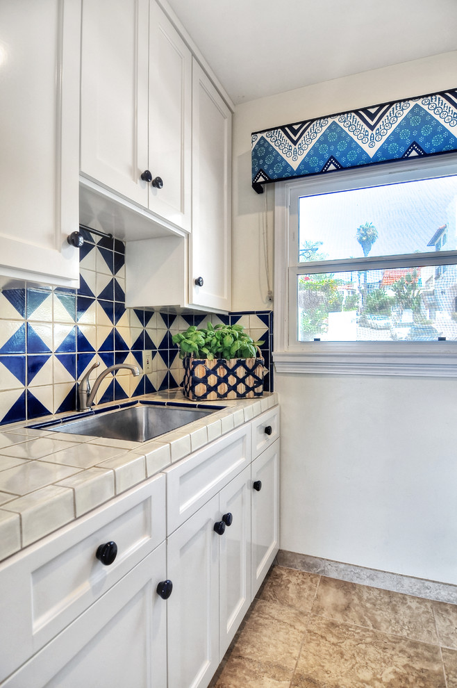 Bild på ett avskilt, litet medelhavsstil vit linjärt vitt kök, med en undermonterad diskho, luckor med upphöjd panel, vita skåp, kaklad bänkskiva, blått stänkskydd, stänkskydd i keramik, rostfria vitvaror och klinkergolv i porslin