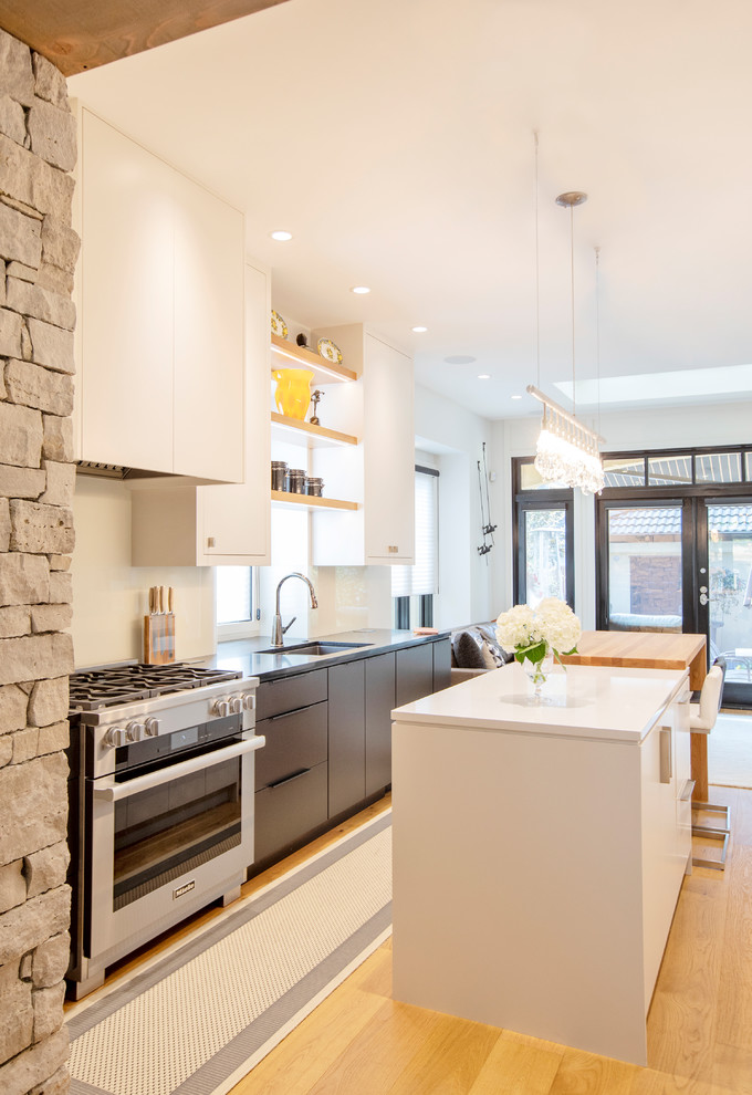 Example of a mid-sized trendy galley medium tone wood floor and white floor kitchen design in Vancouver with an undermount sink, flat-panel cabinets, white cabinets, stainless steel appliances, an island, white countertops, quartz countertops and glass sheet backsplash