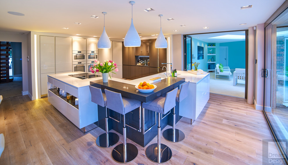 This is an example of a large contemporary l-shaped kitchen/diner in Dorset with a double-bowl sink, flat-panel cabinets, white cabinets, composite countertops, white appliances, porcelain flooring and a breakfast bar.