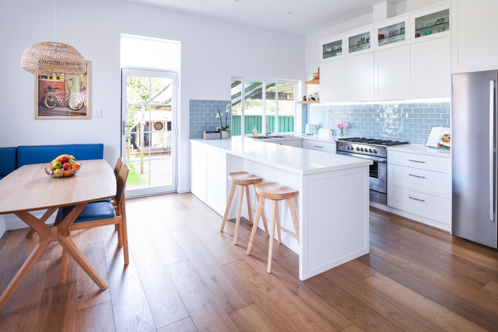 Bild på ett funkis vit vitt kök, med skåp i shakerstil, vita skåp, blått stänkskydd, stänkskydd i tunnelbanekakel, rostfria vitvaror, mellanmörkt trägolv, en halv köksö och brunt golv