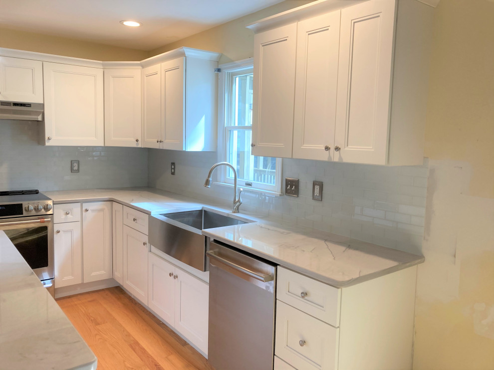 Mid-sized minimalist l-shaped medium tone wood floor and brown floor enclosed kitchen photo in Philadelphia with a farmhouse sink, shaker cabinets, white cabinets, granite countertops, gray backsplash, glass tile backsplash, stainless steel appliances, an island and gray countertops