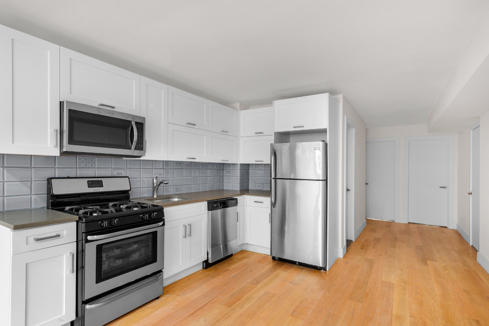 Photo of a small modern l-shaped kitchen/diner in New York with shaker cabinets, no island, a submerged sink, white cabinets, composite countertops, blue splashback, ceramic splashback, stainless steel appliances, light hardwood flooring and beige floors.