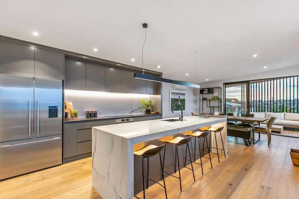 Contemporary galley kitchen/diner in Auckland with a submerged sink, flat-panel cabinets, grey cabinets, grey splashback, stainless steel appliances, medium hardwood flooring, an island, brown floors and grey worktops.