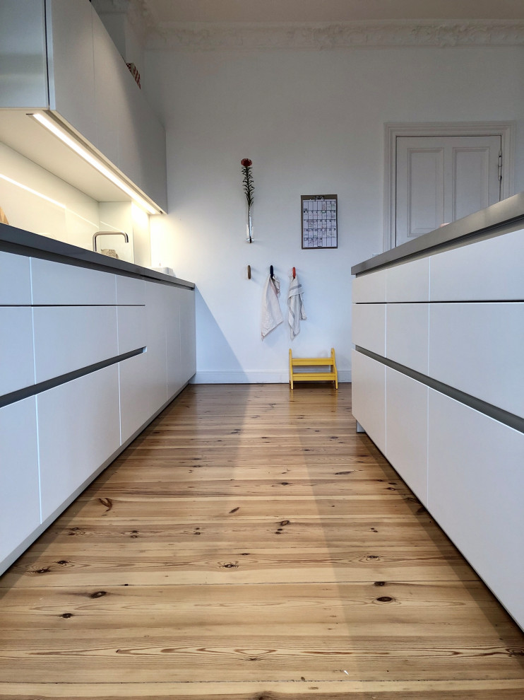 Inredning av ett modernt stort grå linjärt grått kök och matrum, med en nedsänkt diskho, släta luckor, vita skåp, bänkskiva i kalksten, vitt stänkskydd, glaspanel som stänkskydd, svarta vitvaror, ljust trägolv, en köksö och brunt golv