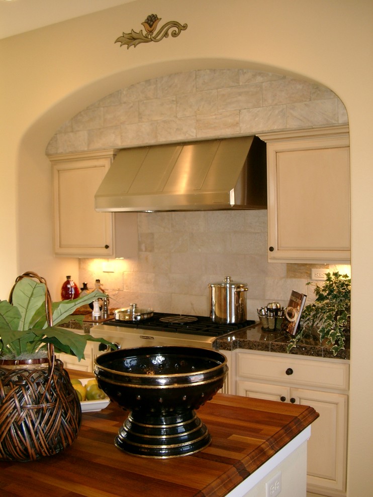 Inspiration for a mediterranean l-shaped eat-in kitchen remodel in Denver with an undermount sink, raised-panel cabinets, white cabinets, beige backsplash, stone tile backsplash, stainless steel appliances, wood countertops and an island