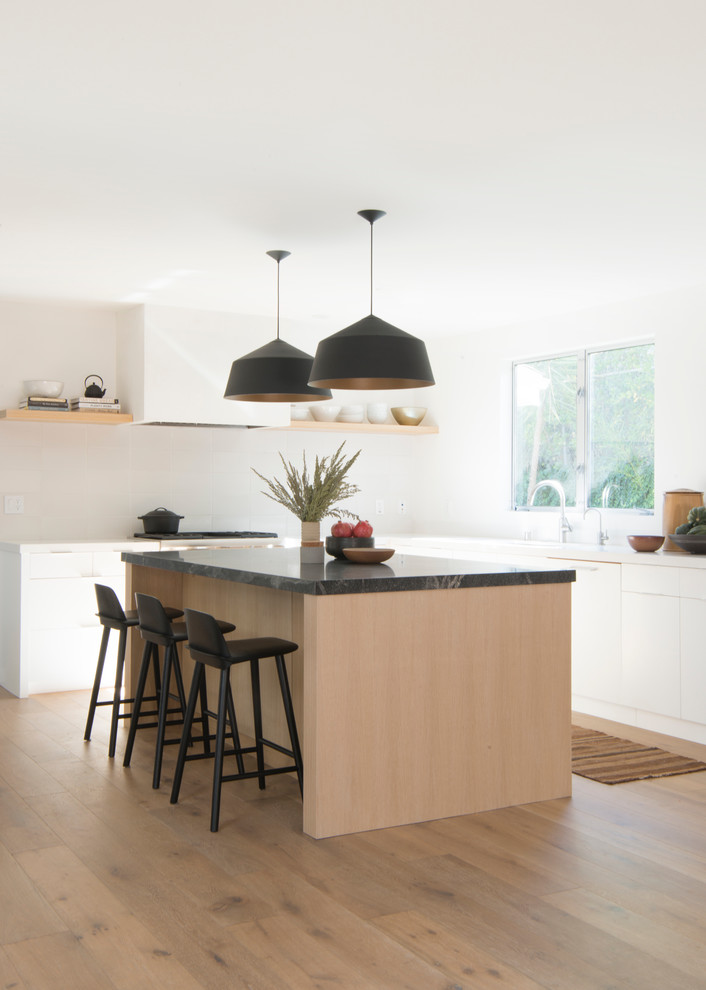 Contemporary kitchen/diner in Los Angeles with a submerged sink, flat-panel cabinets, white cabinets, soapstone worktops, white splashback, ceramic splashback, light hardwood flooring and an island.