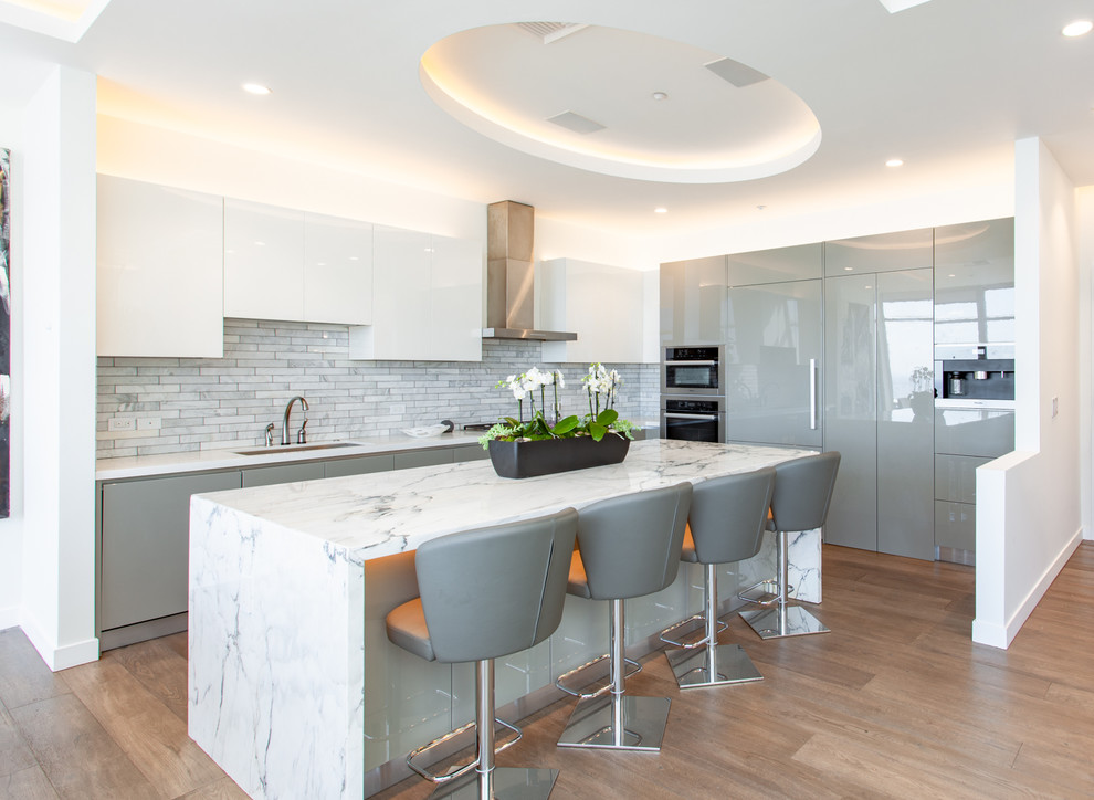 Design ideas for a contemporary l-shaped kitchen in Los Angeles with a submerged sink, flat-panel cabinets, grey cabinets, grey splashback, integrated appliances, medium hardwood flooring, an island, brown floors and white worktops.