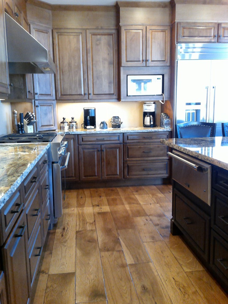 This is an example of a medium sized rustic kitchen/diner in Denver with a belfast sink, raised-panel cabinets, dark wood cabinets, granite worktops, stainless steel appliances, medium hardwood flooring and an island.