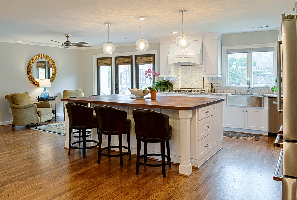 Inspiration for a large cottage l-shaped medium tone wood floor and brown floor open concept kitchen remodel in Charlotte with a farmhouse sink, shaker cabinets, white cabinets, wood countertops, white backsplash, porcelain backsplash, stainless steel appliances and an island