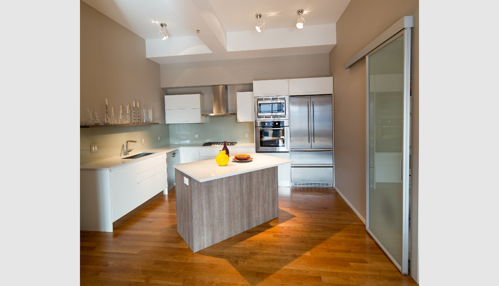 Mid-sized trendy l-shaped medium tone wood floor eat-in kitchen photo in Boston with a drop-in sink, flat-panel cabinets, white cabinets, solid surface countertops, beige backsplash, glass sheet backsplash and stainless steel appliances
