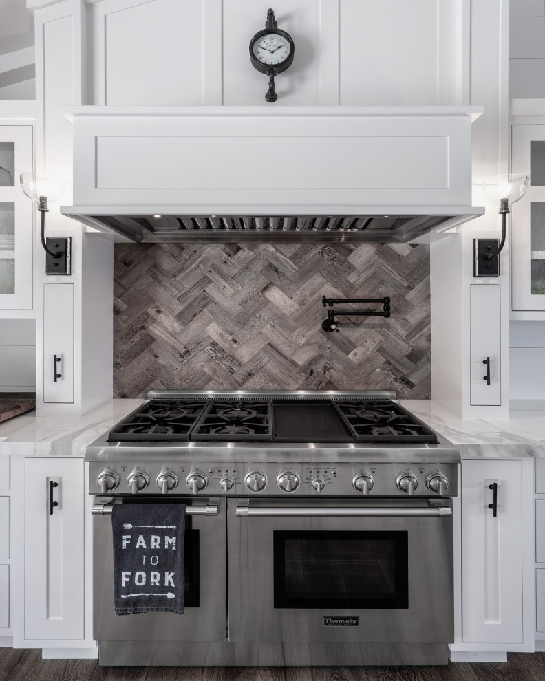 Foto de cocina campestre, de tamaño medio, con fregadero sobremueble, armarios estilo shaker, puertas de armario blancas, salpicadero blanco, salpicadero de madera, electrodomésticos de acero inoxidable, suelo de madera en tonos medios, una isla, suelo marrón, encimeras blancas y encimera de mármol