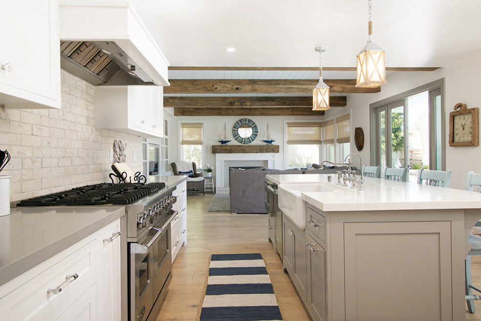 Photo of a large coastal l-shaped kitchen/diner in Orange County with a belfast sink, shaker cabinets, white cabinets, beige splashback, stainless steel appliances, light hardwood flooring, an island, brown floors, engineered stone countertops, stone tiled splashback and grey worktops.