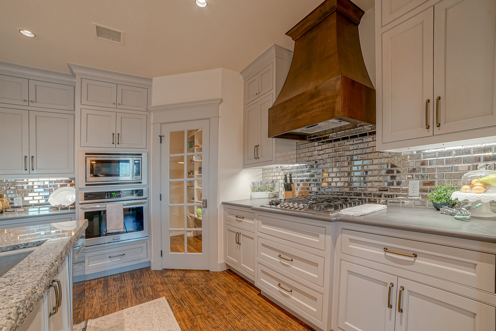 Klassisk inredning av ett stort kök och matrum, med luckor med profilerade fronter, vita skåp, stänkskydd med metallisk yta, spegel som stänkskydd, rostfria vitvaror och en köksö