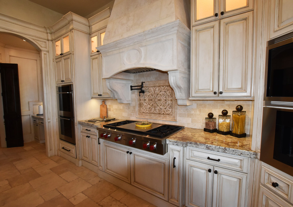 Modelo de cocinas en L mediterránea, extra grande, con despensa, fregadero bajoencimera, armarios con paneles con relieve, puertas de armario con efecto envejecido, encimera de granito, salpicadero beige, salpicadero de azulejos de piedra, electrodomésticos con paneles, suelo de travertino y dos o más islas