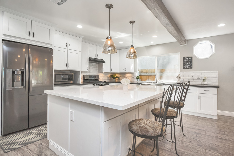 Photo of a medium sized rural l-shaped open plan kitchen in Orange County with a belfast sink, shaker cabinets, white cabinets, engineered stone countertops, white splashback, metro tiled splashback, stainless steel appliances, laminate floors and an island.