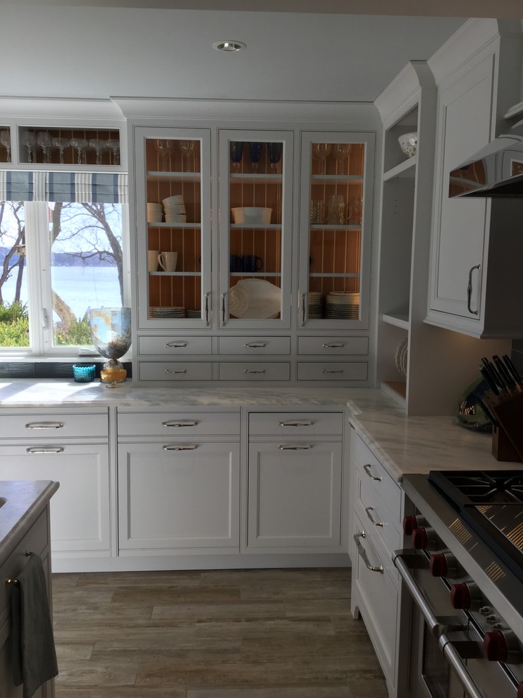 Inspiration for a medium sized nautical l-shaped kitchen/diner in Milwaukee with a belfast sink, beaded cabinets, white cabinets, marble worktops, blue splashback, glass tiled splashback, stainless steel appliances, ceramic flooring, an island, brown floors and blue worktops.