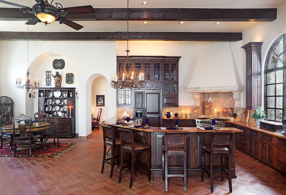На фото: кухня-гостиная в средиземноморском стиле с темными деревянными фасадами, стеклянными фасадами, техникой под мебельный фасад и барной стойкой с