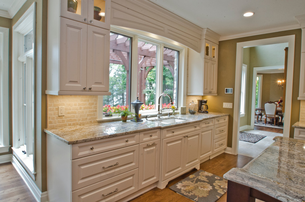 Eat-in kitchen - large traditional medium tone wood floor eat-in kitchen idea in Milwaukee with a drop-in sink, raised-panel cabinets, white cabinets, granite countertops, beige backsplash, subway tile backsplash, stainless steel appliances and an island