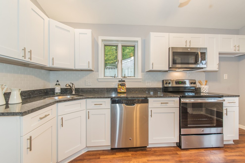 Example of a mid-sized transitional l-shaped medium tone wood floor and brown floor enclosed kitchen design in New York with a drop-in sink, shaker cabinets, white cabinets, granite countertops, white backsplash, ceramic backsplash and stainless steel appliances