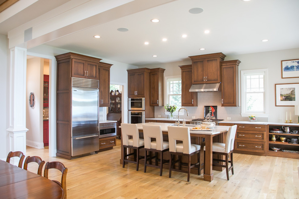 This is an example of a large traditional l-shaped kitchen/diner in Milwaukee with recessed-panel cabinets, dark wood cabinets, engineered stone countertops, white splashback, metro tiled splashback, an island, white worktops, a submerged sink, stainless steel appliances, light hardwood flooring and beige floors.