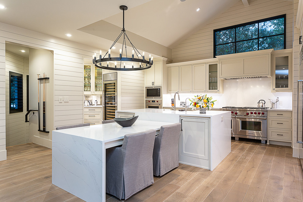 Inspiration for a large farmhouse l-shaped medium tone wood floor and brown floor enclosed kitchen remodel in Austin with a farmhouse sink, shaker cabinets, beige cabinets, marble countertops, stainless steel appliances, an island and white countertops