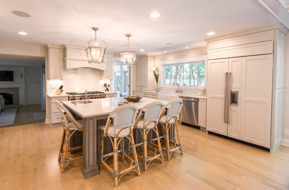 This is an example of a large traditional u-shaped enclosed kitchen in Milwaukee with a belfast sink, recessed-panel cabinets, porcelain splashback, stainless steel appliances, light hardwood flooring and an island.