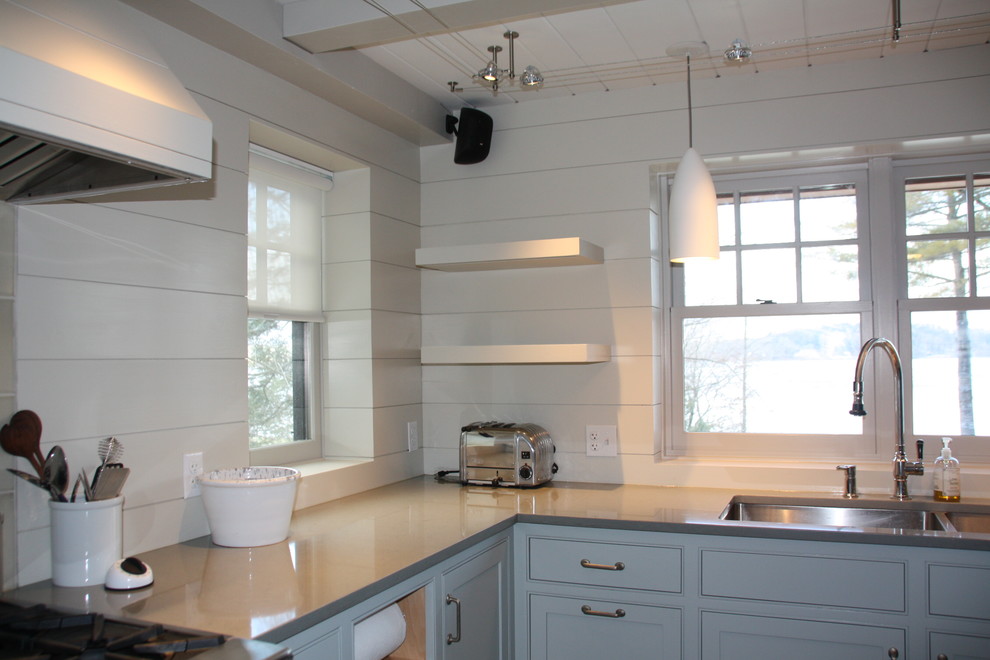 Bild på ett funkis l-kök, med en dubbel diskho, skåp i shakerstil, blå skåp, bänkskiva i kvarts, vitt stänkskydd, stänkskydd i trä, rostfria vitvaror, ljust trägolv, en köksö och blått golv
