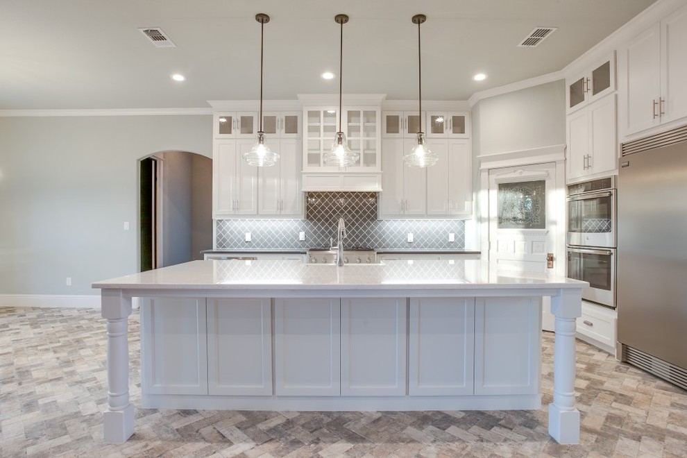 Large traditional l-shaped open plan kitchen in Dallas with a belfast sink, shaker cabinets, white cabinets, granite worktops, grey splashback, glass tiled splashback, stainless steel appliances, brick flooring, an island and grey floors.