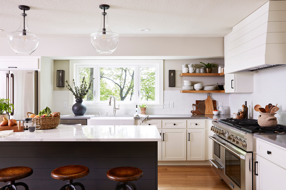 Design ideas for a medium sized beach style l-shaped kitchen/diner in Minneapolis with a belfast sink, shaker cabinets, white cabinets, engineered stone countertops, white splashback, ceramic splashback, stainless steel appliances, medium hardwood flooring, an island and white worktops.