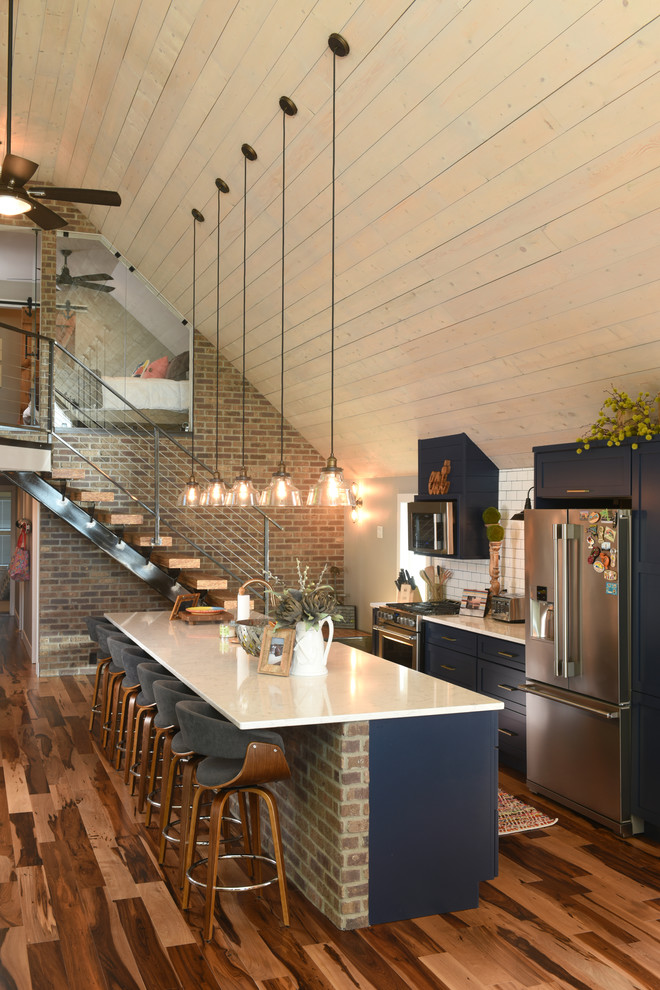 This is an example of a rustic galley kitchen in Other with shaker cabinets, blue cabinets, white splashback, metro tiled splashback, stainless steel appliances, medium hardwood flooring, an island, brown floors and white worktops.