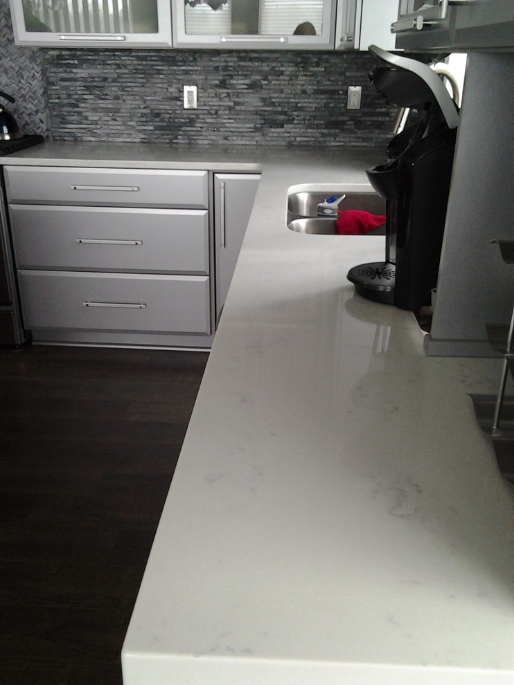 Bild på ett mellanstort funkis vit vitt kök, med en undermonterad diskho, släta luckor, skåp i rostfritt stål, bänkskiva i kvartsit, grått stänkskydd, stänkskydd i stenkakel, rostfria vitvaror, mellanmörkt trägolv, en köksö och grått golv
