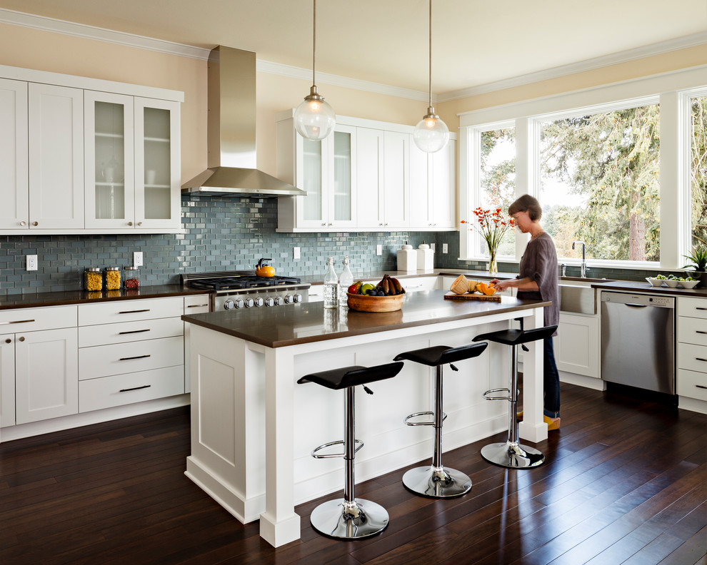 Example of a mid-sized transitional galley dark wood floor kitchen design in Portland with a farmhouse sink, shaker cabinets, white cabinets, mosaic tile backsplash, stainless steel appliances, an island, brown countertops, quartz countertops and brown backsplash