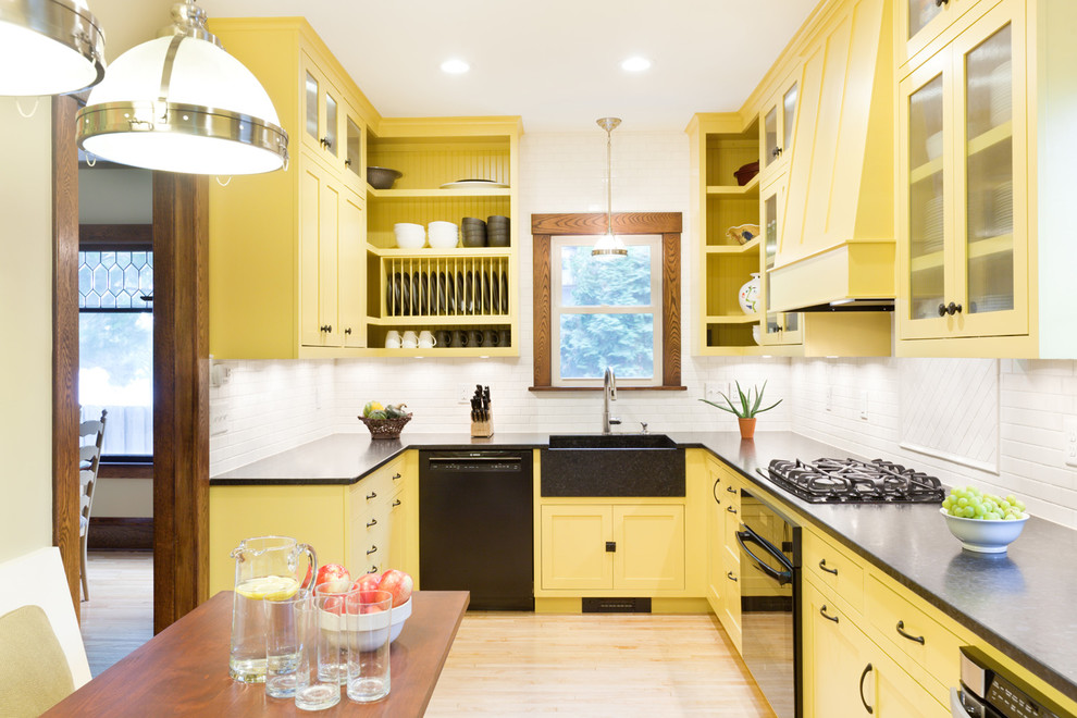 Mittelgroße Klassische Wohnküche ohne Insel in L-Form mit Landhausspüle, gelben Schränken, Granit-Arbeitsplatte, Küchenrückwand in Weiß, Rückwand aus Keramikfliesen, schwarzen Elektrogeräten, hellem Holzboden und Schrankfronten im Shaker-Stil in Minneapolis