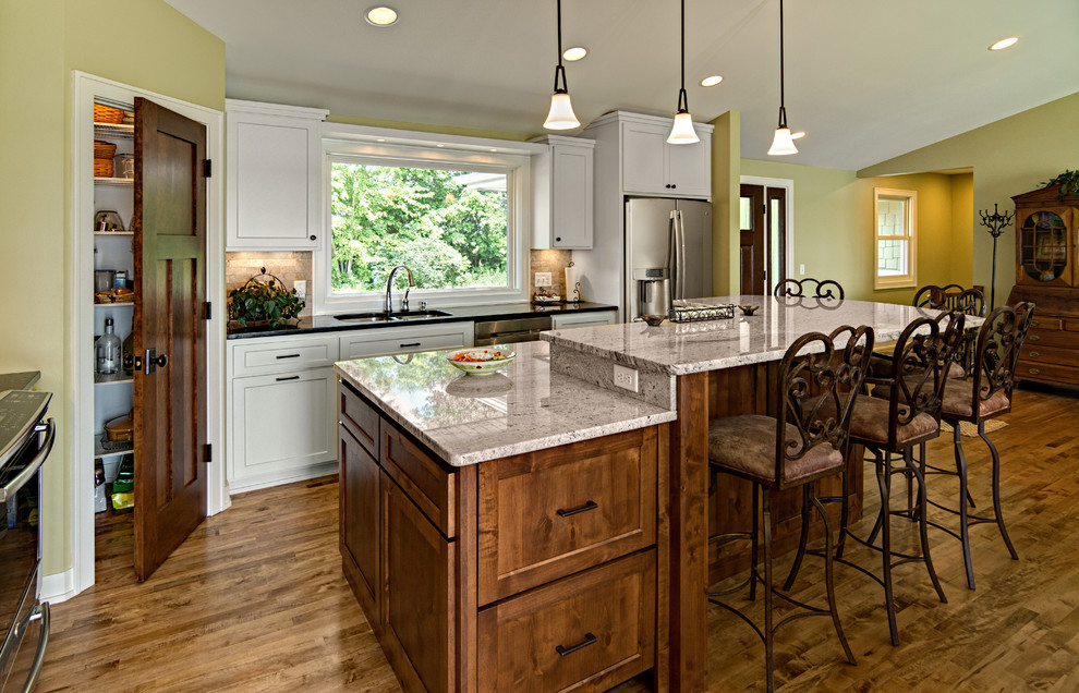 This is an example of a medium sized traditional l-shaped open plan kitchen in Minneapolis with a double-bowl sink, shaker cabinets, dark wood cabinets, granite worktops, beige splashback, stone tiled splashback, stainless steel appliances, medium hardwood flooring, an island, brown floors and multicoloured worktops.