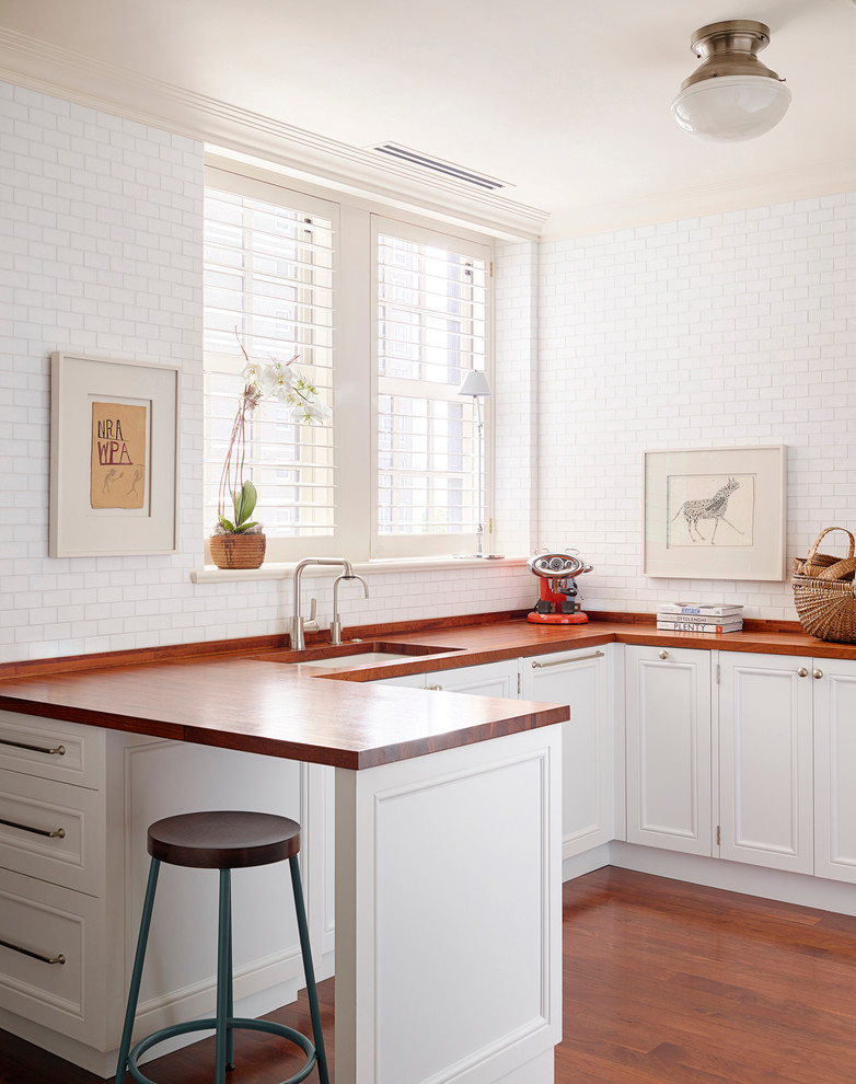 Klassisk inredning av ett u-kök, med en undermonterad diskho, luckor med infälld panel, vita skåp, träbänkskiva, vitt stänkskydd, stänkskydd i tunnelbanekakel, mellanmörkt trägolv, en halv köksö och integrerade vitvaror