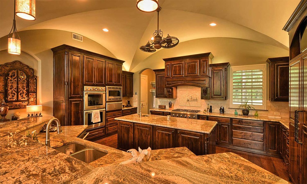 Inspiration for a large timeless u-shaped medium tone wood floor kitchen pantry remodel in Dallas with an undermount sink, raised-panel cabinets, dark wood cabinets, granite countertops, beige backsplash, stone tile backsplash, stainless steel appliances and two islands