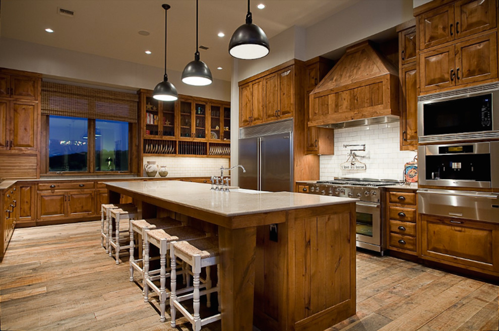 Example of a large mountain style medium tone wood floor kitchen design in Austin with a farmhouse sink, recessed-panel cabinets, medium tone wood cabinets, granite countertops, beige backsplash, subway tile backsplash, stainless steel appliances and an island