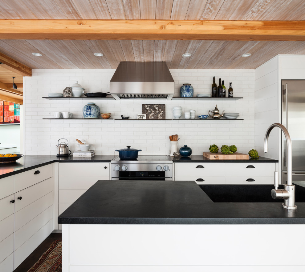 Beach style l-shaped exposed beam and wood ceiling kitchen photo in Seattle with a farmhouse sink, flat-panel cabinets, white cabinets, white backsplash, subway tile backsplash, stainless steel appliances, an island and black countertops
