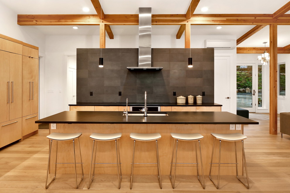 Photo of a contemporary l-shaped open plan kitchen in Seattle with a built-in sink, flat-panel cabinets, light wood cabinets, granite worktops, black splashback, stone tiled splashback, light hardwood flooring, an island, brown floors, black worktops and integrated appliances.