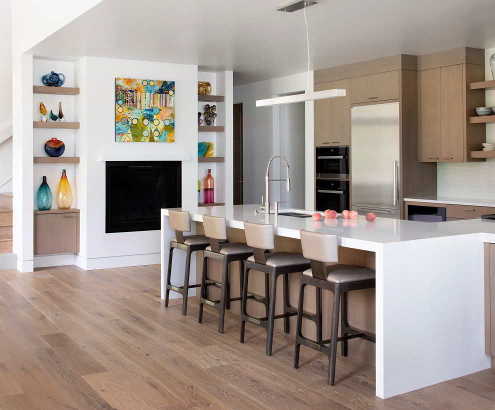 Moderne Küche mit Unterbauwaschbecken, flächenbündigen Schrankfronten, hellbraunen Holzschränken, Küchenrückwand in Weiß, Küchengeräten aus Edelstahl, braunem Holzboden, Kücheninsel, braunem Boden und weißer Arbeitsplatte in Sonstige