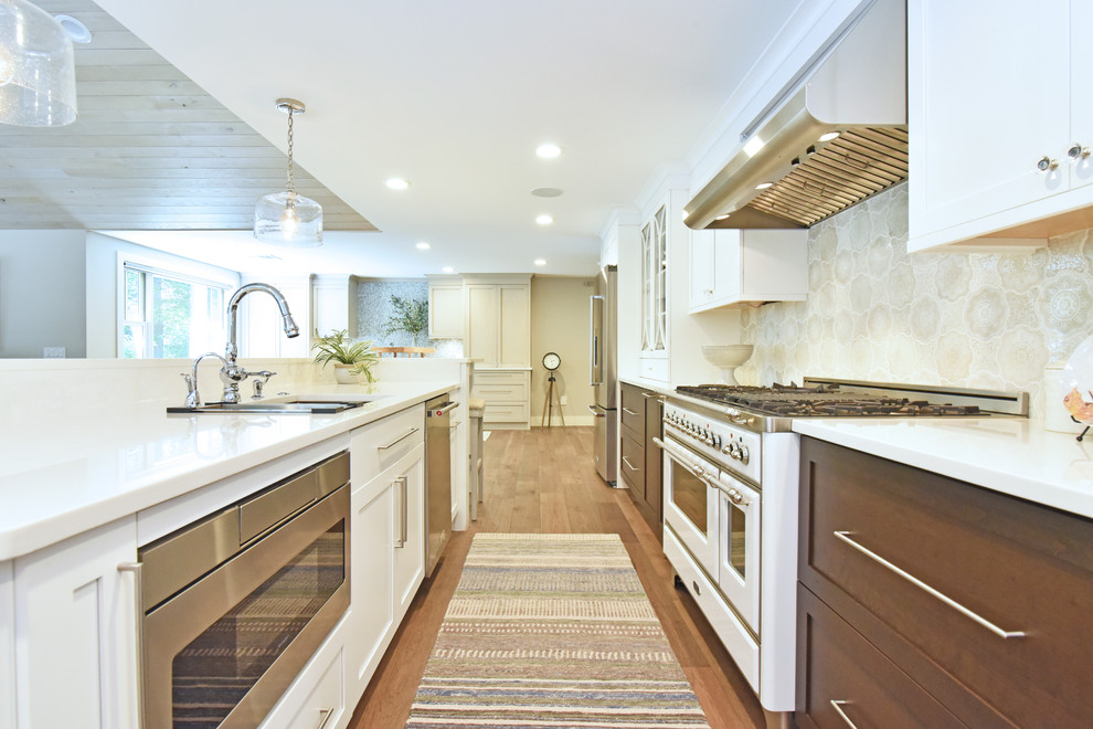 Klassisk inredning av ett mellanstort vit vitt kök, med en undermonterad diskho, luckor med infälld panel, vita skåp, rostfria vitvaror, mörkt trägolv, brunt golv, bänkskiva i kvarts, grått stänkskydd, stänkskydd i keramik och en köksö