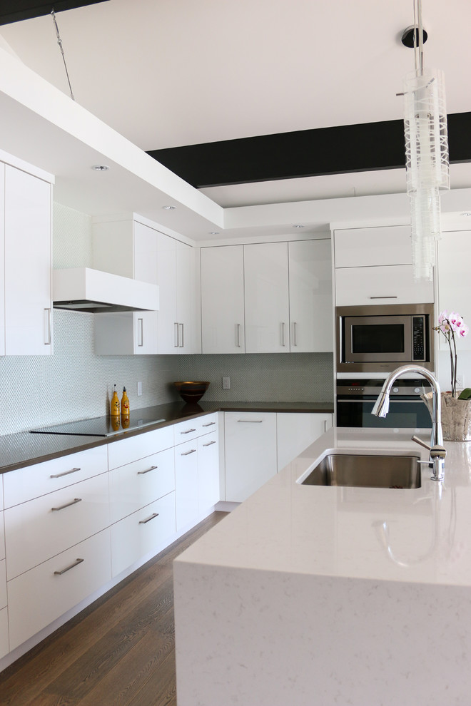 Mittelgroße Moderne Küche in L-Form mit Unterbauwaschbecken, flächenbündigen Schrankfronten, weißen Schränken, Quarzwerkstein-Arbeitsplatte, Rückwand aus Glasfliesen, Küchengeräten aus Edelstahl, braunem Holzboden und Kücheninsel in Vancouver