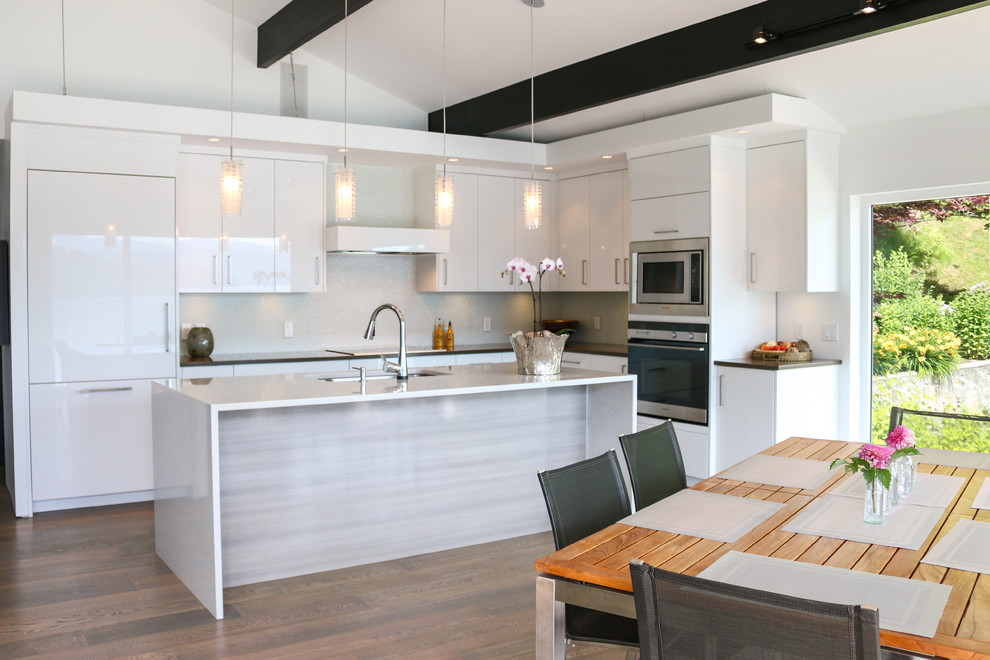 Mittelgroße Moderne Küche in L-Form mit flächenbündigen Schrankfronten, weißen Schränken, Quarzwerkstein-Arbeitsplatte, Kücheninsel, Unterbauwaschbecken, Küchengeräten aus Edelstahl, braunem Holzboden und Rückwand aus Glasfliesen in Vancouver