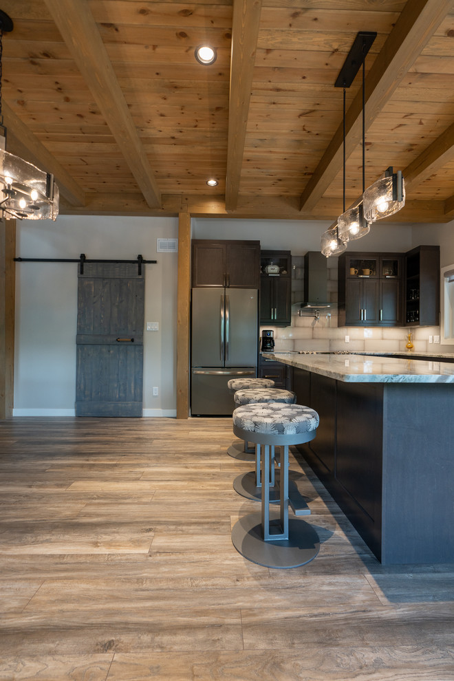 Example of a mid-sized mountain style l-shaped medium tone wood floor and brown floor open concept kitchen design with an undermount sink, shaker cabinets, black cabinets, beige backsplash, stainless steel appliances, an island and multicolored countertops